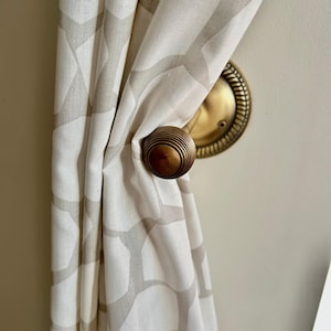 Könnte beinhalten: Ein Vorhang-Halter aus Messing mit einem dekorativen Knopf und einer runden Basis. Der Halter hält einen weißen Vorhang mit geometrischem Muster.