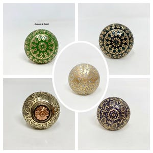 May include: A selection of decorative cabinet knobs in different colours and patterns. The knobs display detailed designs in gold, green, black, and navy blue. One knob is gold with a floral pattern, and another has a copper-coloured centre.