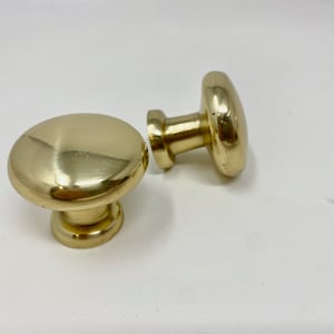 以下が含まれることがあります： 丸みを帯びたキノコ型のデザインの、磨かれた真鍮製のキャビネットノブが2つ。ノブは暖かく金色の色合いで、滑らかで反射する表面を持っています。1つのノブは直立しており、もう1つは横に倒れており、金具のシンプルなエレガンスを示しています。