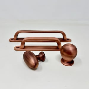 Könnte beinhalten: Ein Satz aus vier Kupfer-Schrankbeschlägen. Zwei sind gebogene Griffe, einer ist ein runder Knopf und einer ist ein ovaler Knopf.