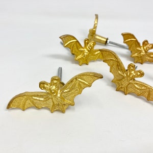May include: Four gold-coloured bat-shaped cabinet knobs with silver screws. The bats have detailed wings and bodies.