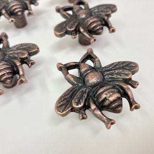 Może przedstawiać: Zestaw pięciu antycznych, miedzianych gałek do szuflad w kształcie pszczoły. Projekt pszczoły jest szczegółowy i ma postarzaną powierzchnię.