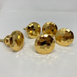 Può includere: Quattro pomelli per armadi di colore dorato con finitura martellata. I pomelli sono rotondi e hanno un centro leggermente rialzato.