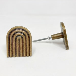 Op de afbeelding: Twee gouden kastknoppen met een regenboogvormig ontwerp. Eén knop is bevestigd aan een schroef, de andere niet.