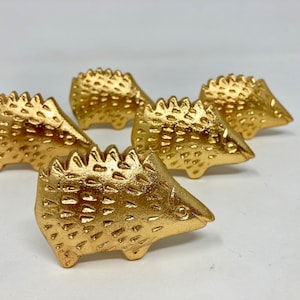 Op de afbeelding: Vijf gouden keramische egelbeeldjes. Elk beeldje heeft een getextureerd oppervlak en een gedetailleerd gezicht.