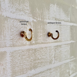 Peut inclure: Deux crochets en laiton sur un mur de briques blanches. Le crochet de gauche est en laiton poli. Le crochet de droite est en laiton antique.