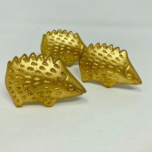 Op de afbeelding: Drie gouden egelbeeldjes. De egels zijn gedetailleerd met getextureerde stekels en hebben een realistische vorm.