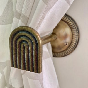 May include: A gold-toned metal curtain tieback with a decorative arched design. The tieback is attached to a wall-mounted bracket.