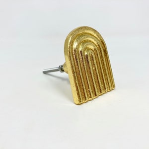 May include: Gold-toned cabinet knob with a rounded rectangular shape and a textured design resembling an archway.