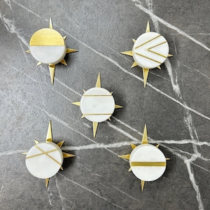 May include: Five decorative compass-shaped objects. Each has a white, circular base with gold accents and a gold starburst design. The gold accents include stripes and a half-circle. They are arranged on a grey marble surface.