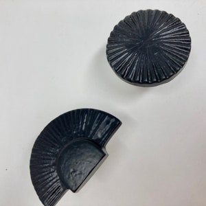 Könnte beinhalten: Zwei schwarze Keramik-Schrankknöpfe mit Sonnenstrahlen-Design. Ein Knopf ist rund und der andere ist ein Halbkreis.