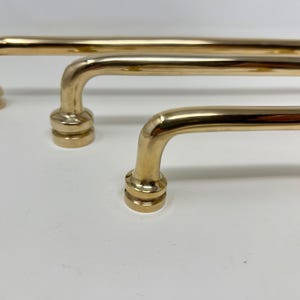 Puede incluir: Tres tiradores de armario de color dorado con un diseño redondeado y ligeramente curvado. Los tiradores están dispuestos en fila, con el tirador más grande en la parte inferior y el más pequeño en la parte superior.