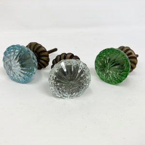 May include: Three decorative glass cabinet knobs in shades of blue, clear, and green. Each knob has a textured, cut-glass design and is attached to a dark bronze-coloured metal base. The knobs are arranged on a white surface.