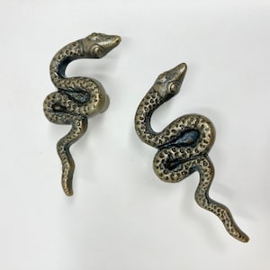 May include: Two antique gold-toned snake-shaped cabinet knobs. The snakes are curled in a realistic pose with detailed scales and eyes.