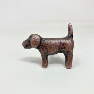 May include: A small, copper-coloured dog figurine with a textured finish. The dog is standing on all four legs with its tail curled up.