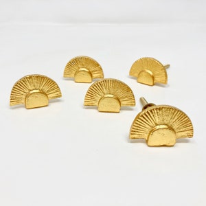 May include: Five gold-toned cabinet knobs with a sunburst design. Each knob has a raised, textured surface and a rounded, half-circle shape.