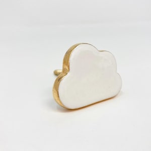 May include: A white cloud-shaped cabinet knob with a gold-colored rim and stem. The knob is made of ceramic or porcelain and is designed for use on drawers or cabinets. The cloud design adds a whimsical touch.