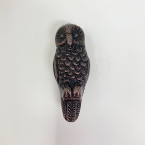 May include: A bronze-colored owl figurine. The owl is depicted in a vertical position, with detailed feather patterns and a textured surface. The owl's eyes and beak are clearly defined, and the overall design is symmetrical.