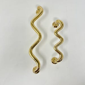 Può includere: Due maniglie per porte in metallo ondulato color oro. Le maniglie sono di dimensioni diverse, la più grande è a sinistra e la più piccola a destra.