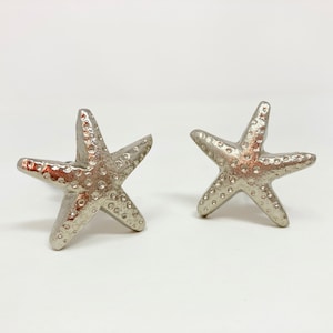 May include: Two silver starfish-shaped drawer pulls. The starfish have a textured surface and are a popular choice for coastal-themed decor.