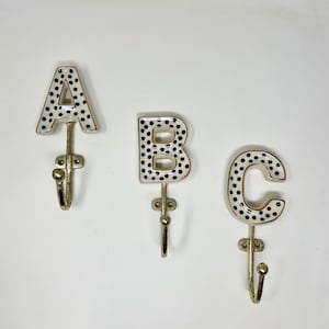 May include: Three decorative wall hooks shaped like the letters A, B, and C. Each letter is white with black polka dots and a gold trim. The hooks are made of metal and have a vintage style.