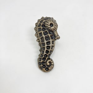 May include: A gold-toned metal seahorse figurine with a detailed design. The seahorse has a curled tail and a textured body.