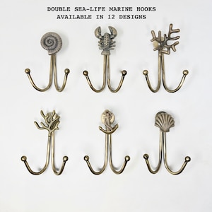 Puede incluir: Seis ganchos dobles de latón antiguo con diseños marinos, incluyendo una concha, una langosta y coral. El texto en la parte superior dice "DOUBLE SEA-LIFE MARINE HOOKS AVAILABLE IN 12 DESIGNS."