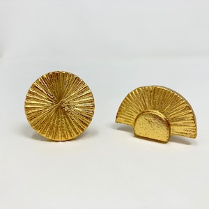 May include: Two gold-toned metal drawer pulls. One is a round knob with a sunburst design. The other is a half-circle shape with a sunburst design.