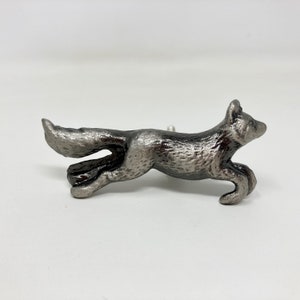 Pode incluir: Uma figura de raposa de metal prateado em uma pose de corrida. A raposa tem uma superfície detalhada e texturizada e é um acento decorativo para uma casa.
