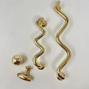 Könnte beinhalten: Eine Sammlung goldfarbener Beschläge, darunter zwei wellenförmige Griffe, ein runder Knauf und ein kleiner Ziergriff. Die Beschläge haben eine polierte Oberfläche, die das Licht reflektiert. Die Griffe haben ein einzigartiges, serpentinenförmiges Design.