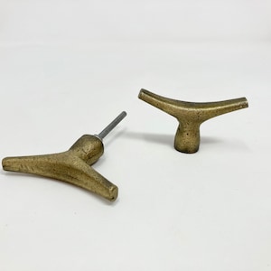 Puede incluir: Dos tiradores de puerta de armario de color dorado antiguo. Un tirador tiene forma de T con la parte superior redondeada y el otro tiene forma de V con la parte inferior redondeada.