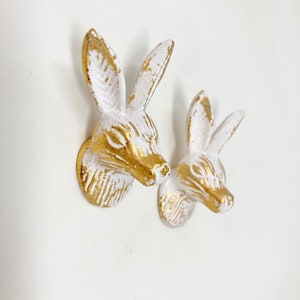 Op de afbeelding: Twee witte keramische wandhaken in de vorm van een konijnenkop met gouden accenten. De haken zijn bevestigd aan een witte muur.