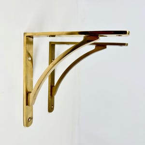 Puede incluir: Dos escuadras de estante de latón. Cada escuadra tiene un soporte vertical plano y rectangular con esquinas redondeadas y un soporte de estante horizontal curvado. Las escuadras son de color dorado cálido y están montadas en una pared blanca.