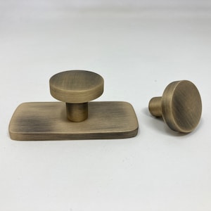 Puede incluir: Dos pomos de armario de color dorado antiguo. Un pomo está unido a una placa base rectangular.