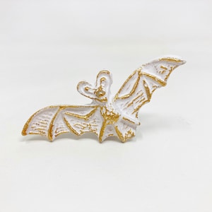Könnte beinhalten: Eine weiße Fledermausfigur mit goldenen Akzenten. Die Fledermaus ist mit ausgebreiteten Flügeln dargestellt, die detaillierte Merkmale aufweisen. Goldene Akzente heben die Kanten der Flügel und des Körpers hervor und erzeugen einen Kontrast.