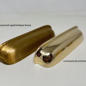 May include: Two unlacquered brass cabinet pulls, one with an aged/antique finish and one with a polished finish. The text "unlacquered aged/antique brass" is below the aged pull and the text "unlacquered polished brass" is below the polished pull.