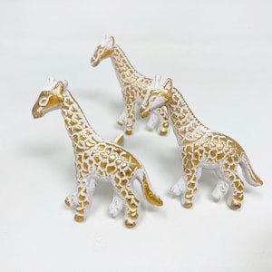 Könnte beinhalten: Drei weiße Keramik-Schubladengriffe in Giraffenform mit goldenen Akzenten. Die Giraffen stehen mit dem Kopf zur Seite gedreht.