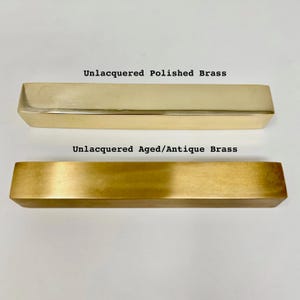 May include: Two rectangular pieces of unlacquered brass. The top piece is polished and shiny. The bottom piece is aged and has a more matte finish. The text "Unlacquered Polished Brass" is above the top piece and the text "Unlacquered Aged/Antique Brass" is above the bottom piece.