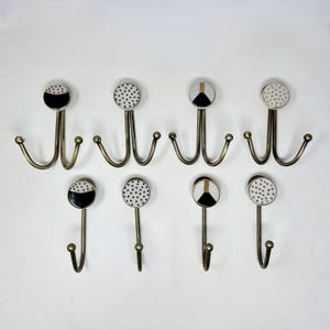 May include: Eight decorative wall hooks with brass-colored metal hooks and ceramic tops. The ceramic tops feature black and white geometric patterns, including polka dots and triangles. The hooks are arranged in two rows of four.