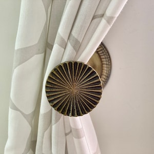 May include: Close-up of a decorative curtain tieback. The tieback is a circular, bronze-coloured metal piece with a sunburst design. The curtain is white with a grey geometric pattern.