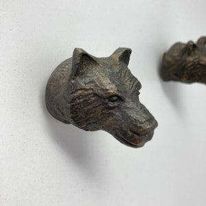 Peut inclure: Deux crochets muraux en bronze en forme de tête de loup. Les têtes de loup sont tournées vers la droite. Les crochets sont fixés à un mur blanc.