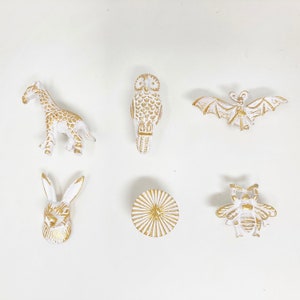 Op de afbeelding: Set van zes witte keramische ladegrepen met gouden accenten. De grepen hebben dierlijke en geometrische ontwerpen, waaronder een giraffe, een uil, een vleermuis, een konijn, een bij en een zonnestralen.