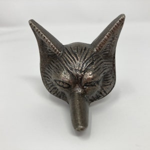 Puede incluir: Un tapón de botella de metal plateado en forma de cabeza de zorro. El zorro tiene una cara detallada con un hocico largo y orejas puntiagudas.