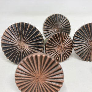 May include: Close-up of several bronze-coloured cabinet knobs. Each knob is round with a sunburst design radiating from the centre. The knobs have a slightly distressed finish, adding to their vintage appeal. The knobs are arranged on a white surface.