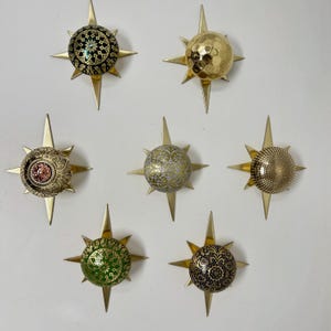 May include: A collection of seven decorative starburst wall art pieces. Each starburst features a central, spherical element with various textures and patterns, including gold, green, and black designs. The starbursts have gold-colored, pointed rays extending outwards.