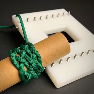 May include: A green paracord bracelet being woven on a white plastic loom with a brown cardboard tube in the center.