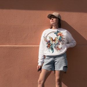 May include: White sweatshirt with a colorful butterfly and floral design on the front. The design features a dreamcatcher with feathers and flowers.