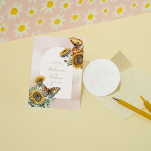 May include: A wedding invitation card featuring a watercolour design of sunflowers and butterflies. The card is pink and white with gold lettering. The text on the card reads "The Wedding of Helene & Pedro 20.08.2023".