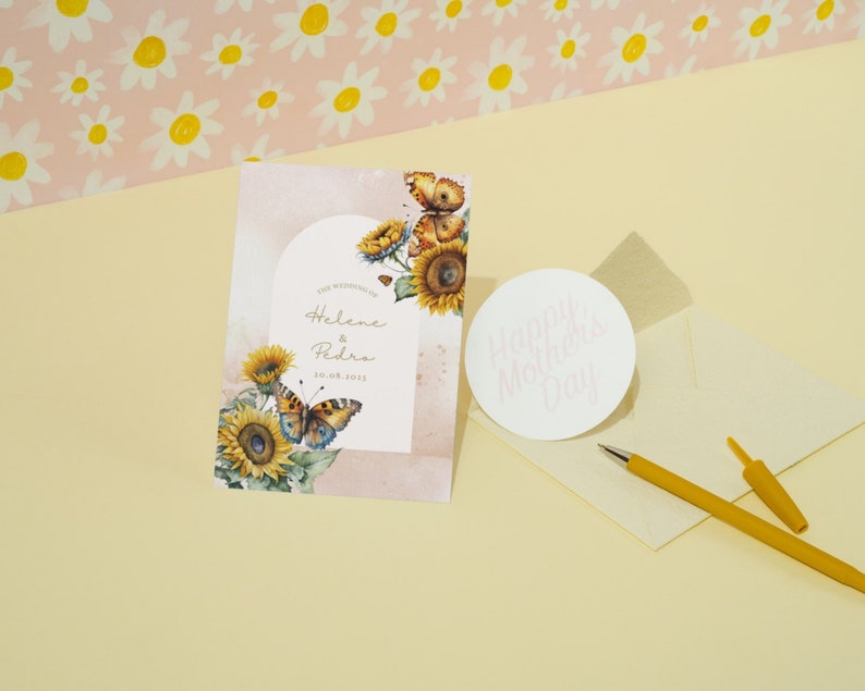 May include: A wedding invitation card featuring a watercolour design of sunflowers and butterflies. The card is pink and white with gold lettering. The text on the card reads "The Wedding of Helene & Pedro 20.08.2023".