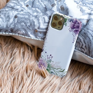 May include: A white phone case with a floral design featuring purple flowers and green leaves. The case has a camera lens cutout and is lying on a brown fuzzy surface.